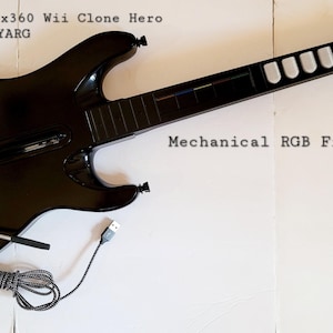 May include: Black guitar-shaped game controller with a long neck and a cable. The text on the image says "PS3 PS4 PS5 Xbox360 Wii Clone Hero YARG" and "Mechanical RGB Frets light up!".
