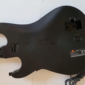 May include: Black Guitar Hero video game controller with a guitar-shaped design. The back of the controller features the Guitar Hero logo and a compartment for batteries. A black and white striped cord extends from the bottom.