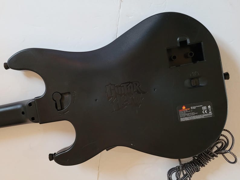 May include: Black Guitar Hero video game controller with a guitar-shaped design. The back of the controller features the Guitar Hero logo and a compartment for batteries. A black and white striped cord extends from the bottom.