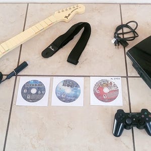 May include: A black and blue Fender guitar controller, a black guitar strap, three game discs, a black PlayStation 3 console, a black game controller, and a power cable. The console features the text PlayStation 3 on its top surface.