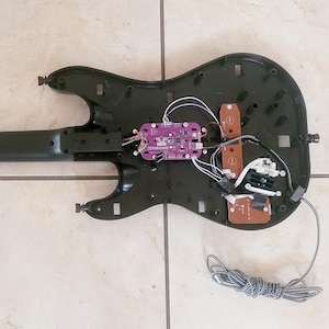 May include: Black guitar-shaped game controller with the back panel removed, revealing internal components. A purple circuit board labeled "ZEROBOUND" is visible, along with wires and other electronic parts. A gray and white striped cable is connected to the controller.