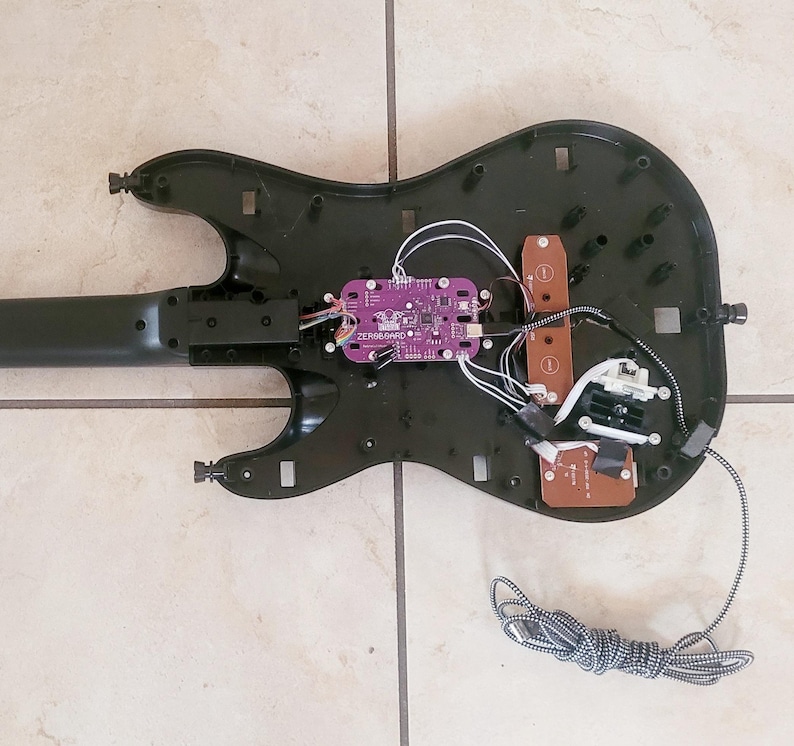 May include: Black guitar-shaped game controller with the back panel removed, revealing internal components. A purple circuit board labeled "ZEROBOUND" is visible, along with wires and other electronic parts. A gray and white striped cable is connected to the controller.