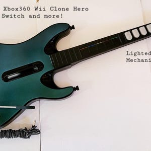 May include: A teal and black guitar-shaped game controller with lighted buttons and mechanical frets. The controller is designed for PS3, PS4, PS5, Xbox360, Wii, and YARG Switch. The image includes the text "Lighted buttons & Mechanical frets."