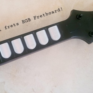 May include: Black guitar controller neck with white fret buttons and the text "Mechanical frets RGB Fretboard!". The headstock has six black tuning pegs. The fretboard has colored lines.