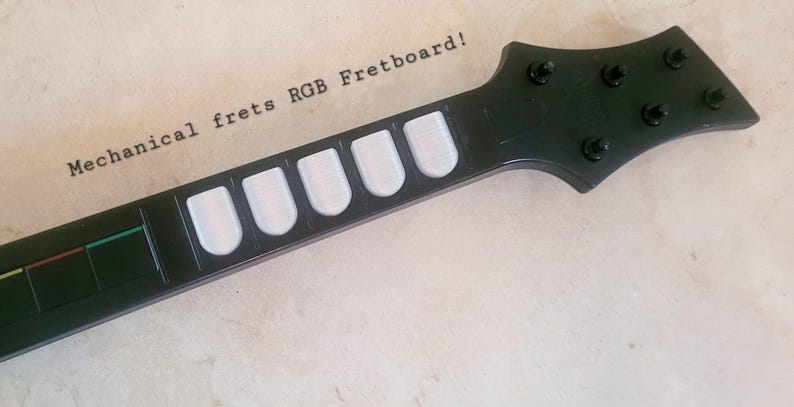 May include: Black guitar controller neck with white fret buttons and the text "Mechanical frets RGB Fretboard!". The headstock has six black tuning pegs. The fretboard has colored lines.