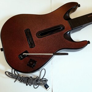 May include: A brown and black toy guitar with a glittery finish. The guitar has a black neck and a silver whammy bar. A black and white striped USB cable is coiled below the guitar.