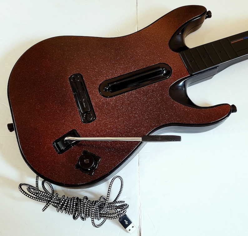 May include: A brown and black toy guitar with a glittery finish. The guitar has a black neck and a silver whammy bar. A black and white striped USB cable is coiled below the guitar.
