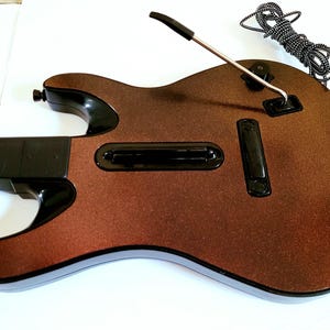 May include: A brown and black guitar-shaped video game controller. The controller has a metallic brown finish with black accents, including the neck, buttons, and strum bar. A silver metal bar and a black and white striped cord extend from the body.
