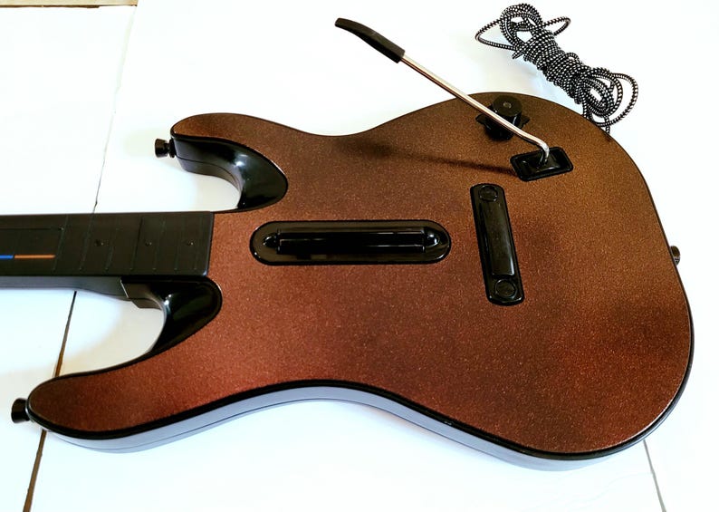 May include: A brown and black guitar-shaped video game controller. The controller has a metallic brown finish with black accents, including the neck, buttons, and strum bar. A silver metal bar and a black and white striped cord extend from the body.