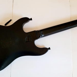 May include: Black guitar-shaped game controller with a long neck and a USB cable. The controller has a matte finish and several buttons and ports. The back of the controller is visible, showing its shape and design.