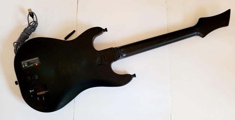 May include: Black guitar-shaped game controller with a long neck and a USB cable. The controller has a matte finish and several buttons and ports. The back of the controller is visible, showing its shape and design.