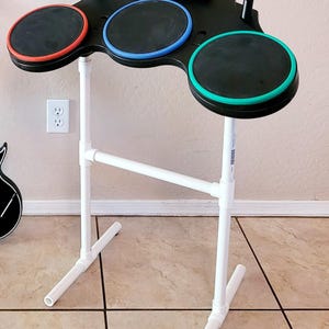 May include: A black electronic drum set stand with three circular pads, each with a different colored rim: red, blue, and green. The stand is white and has the text "Stand Only" at the bottom.