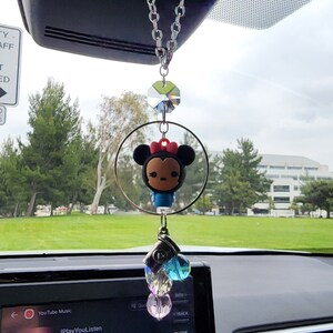 Op de afbeelding: Een auto-hanger met een cartoonfiguur met een rode strik en een kristallen hanger. De hanger hangt aan een zilveren ketting en is bevestigd aan de achteruitkijkspiegel van een auto.