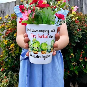 May include: A white ceramic mug with a cactus design and the text "We all bloom uniquely, but in Mrs. Farkas' class we all stick together". A young person is holding the mug in front of a green bush.