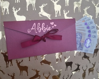 May include: A burgundy envelope with a pink ribbon and the name "Abbie" written on it. The envelope is sitting on a brown background with a pattern of gold reindeer.
