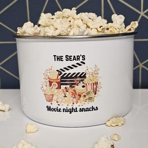 May include: A white enamel mug filled with popcorn, with the text "THE SEAR'S Movie night snacks" printed on the front. The mug has a silver rim and is decorated with movie-themed graphics. Scattered popcorn pieces are in front of the mug.