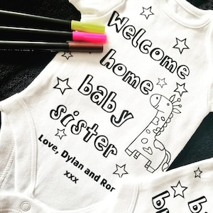 May include: White onesie with black text that reads "Welcome home baby sister" with a black and white giraffe illustration and stars. The text "Love, Dylan and Ror XXX" is also included.