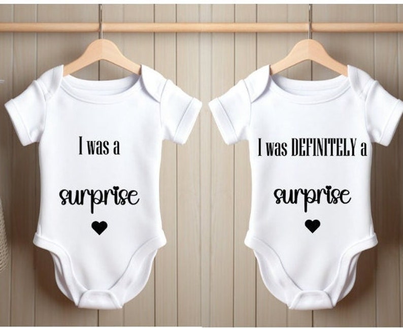 May include: Two white baby onesies with black text. One onesie says "I was a surprise" and the other says "I was definitely a surprise".