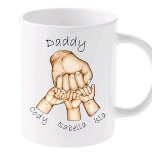 May include: White ceramic mug with a hand-drawn illustration of a father and three children's hands holding each other's fingers. The text "Daddy" is at the top of the mug, and the names "Cody", "Isabella", and "Isla" are written below the hands.