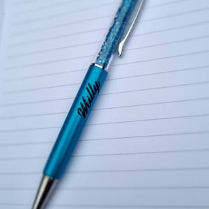 May include: A blue pen with a clear body filled with blue glitter. The pen has a silver clip and a silver stylus tip. The pen is personalized with the name "Milly" in black lettering.