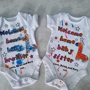 May include: Two white baby onesies with short sleeves. One onesie has a blue giraffe and the text "Welcome home baby brother" with the words "Love, Dylan and Rory xxx". The other onesie has an orange giraffe and the text "Welcome home baby sister" with the words "Love, Dylan and Rory xxx".