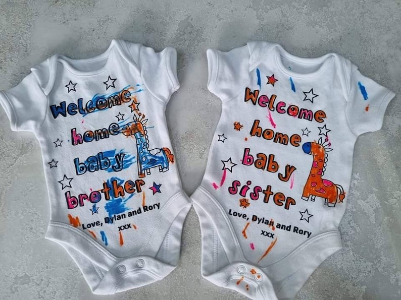 May include: Two white baby onesies with short sleeves. One onesie has a blue giraffe and the text "Welcome home baby brother" with the words "Love, Dylan and Rory xxx". The other onesie has an orange giraffe and the text "Welcome home baby sister" with the words "Love, Dylan and Rory xxx".