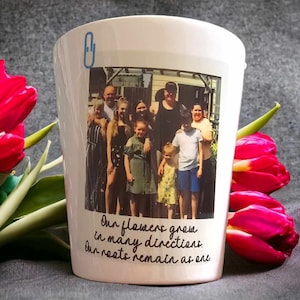 May include: White ceramic mug featuring a photograph of a family of seven standing in front of a house. The photo is framed with a black border and the text "Our flowers grow in many directions. Our roots remain as one." is printed below the image.