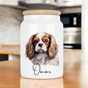 May include: White ceramic jar with a wooden lid, featuring a hand-painted illustration of a Cavalier King Charles Spaniel dog with brown and white fur. The dog's name, "Oscar", is written in cursive below the image.