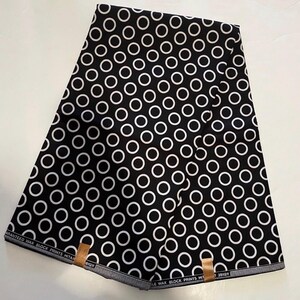 May include: Black fabric featuring a repeating pattern of white circles. The fabric is folded, displaying the pattern across its surface. The edges of the fabric have a striped border with text that reads "GUARANTEED WAX BLOCK PRINTS HITAR".