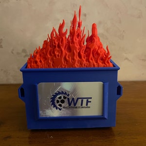 Peut inclure: Une benne à ordures en plastique bleue avec une flamme rouge imprimée en 3D sur le dessus. La benne à ordures a une fenêtre transparente avec un logo qui dit "WIF" et "Weld Technology & Fabrication".