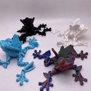 May include: Four articulated frog figurines with butterfly wings. The frogs are in different colors: black, white, blue, and a multi-colored iridescent finish.
