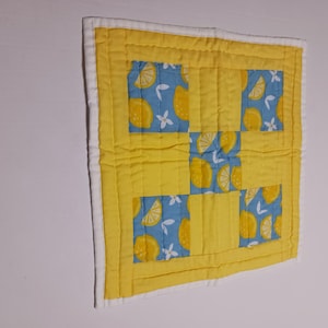 May include: A small, square quilted item with a yellow and blue lemon pattern. The quilt features a yellow border and a white edge. The design includes yellow lemons on a blue background, with white flower accents. The item is likely a decorative piece or a small mat.