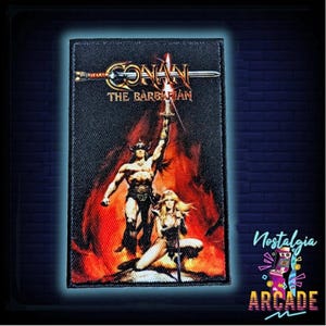 Peut inclure: Écusson rectangulaire avec le texte "CONAN THE BARBARIAN" et une illustration d'un guerrier levant une épée, avec une femme agenouillée en dessous. Le fond représente des flammes. L'écusson a une bordure noire. Le texte "Nostalgia Arcade" est également présent.
