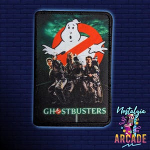 May include: A black and green embroidered patch featuring the Ghostbusters logo and the four Ghostbusters characters standing in front of a green and white background. The logo says "Ghostbusters" in white letters with a red no ghost symbol.