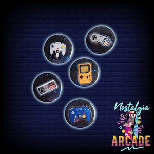 Puede incluir: Cinco botones redondos con un fondo de galaxia e imágenes pixeladas de mandos de videojuegos retro. Los botones están dispuestos en semicírculo con el texto "Nostalgia Arcade" debajo.