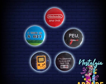 May include: Five round pins with various designs related to video games and gaming. The pins feature a blue pin with the text "I THINK IN & BIT", a red pin with the text "Nintendo and Chill", a black pin with the text "PEW", a black pin with a Game Boy and a black pin with the text "IT'S DANGEROUS TO GO ALONE! TAKE THIS."