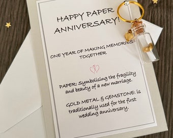 May include: A white card with the text HAPPY PAPER ANNIVERSARY! ONE YEAR OF MAKING MEMORIES TOGETHER, PAPER: Symbolising the fragility and beauty of a new marriage. GOLD METAL & GEMSTONE: is traditionally used for the first wedding anniversary. A glass bottle with a cork and a gold ring attached sits on the card.