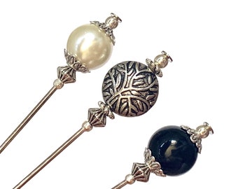 May include: Three decorative hat pins with silver metal accents. The pins have round tops with a white pearl, a black stone, and a silver metal design.