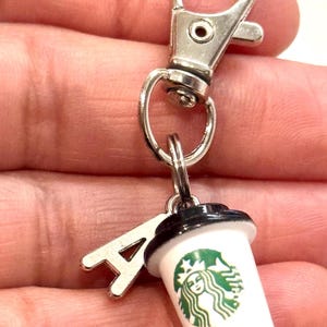 May include: A miniature coffee cup keyring with a silver clasp and a charm shaped like the letter "A". The white cup features the Starbucks logo in green and black. A small, cute accessory.