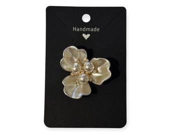 May include: A cream-coloured flower brooch with pearl embellishments and gold-toned details. The brooch is mounted on a black card with the word "Handmade". This is a piece of jewellery.