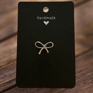 May include: A silver bow-shaped earring on a black card with the word "Handmade" and a heart.