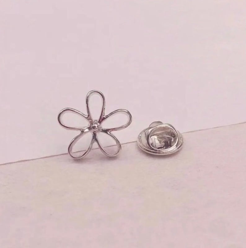 Op de afbeelding: Zilveren speld in de vorm van een bloem met een veiligheidsspeld aan de achterkant. De bloem is een eenvoudig ontwerp met vijf bloemblaadjes.