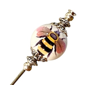 Può includere: Uno spillo da balia d'argento e bianco con una perla rotonda bianca che presenta un'ape dipinta con strisce gialle e nere e ali rosa.