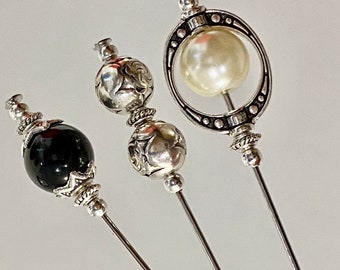 May include: Three silver hat pins with decorative tops. One pin has a black bead, one has a silver bead with an engraved design, and one has a white pearl in a silver circle.