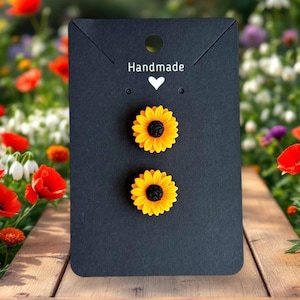 May include: A pair of sunflower earrings with yellow petals and black centres are presented on a black card. The card features the word "Handmade" and a white heart. The background is a blurred garden.