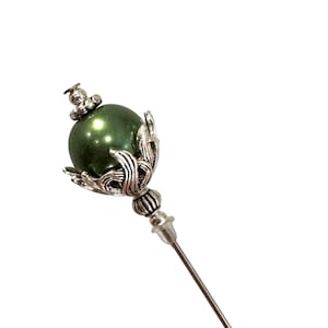 May include: A green glass bead with a silver filigree design and a silver pin. The pin is used for attaching fabric or other materials.