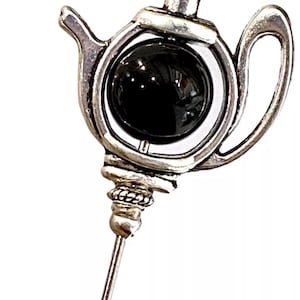 Puede incluir: Pin de plata en forma de tetera con una piedra preciosa negra en el centro. El pin tiene un tallo largo y delgado.