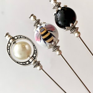 May include: Three silver-toned hat pins with decorative tops. The pins have different coloured glass beads: black, white with a bee design, and a white pearl.