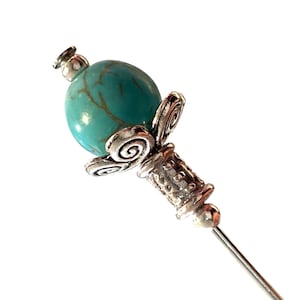 May include: A decorative hat pin with a turquoise bead and silver-toned metal detailing. The pin has a detailed design with a spiral motif and a long, slender pin.
