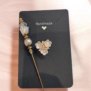 May include: A gold-toned stick pin featuring a clear crystal and white bead topper, accompanied by a three-flower brooch with white petals and small clear rhinestones. Both items are attached to a black rectangular display card with the text Handmade.